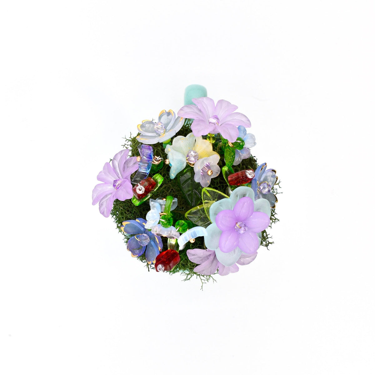 A circular decorative ornament featuring a dense cluster of artificial flowers in shades of lavender, light blue, and white, interspersed with green moss, small translucent glass beads, and tiny red accents.