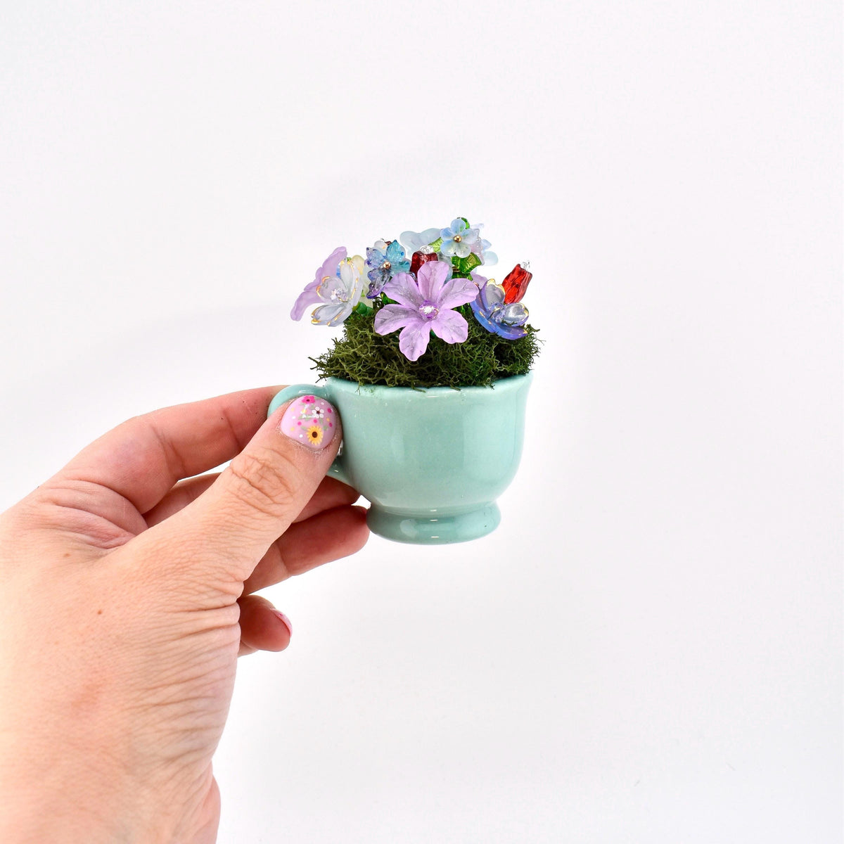 A miniature light blue ceramic teacup filled with green moss and a cluster of small, translucent, multi-colored glass flowers in shades of purple, blue, and red, creating a delicate floral arrangement in a tiny vessel.