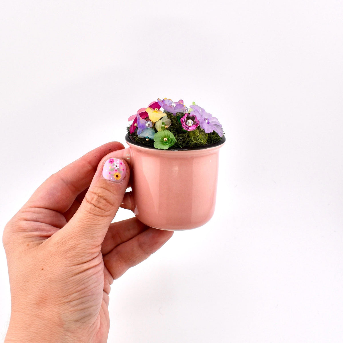A small, light pink ceramic mug filled with green moss and a cluster of miniature, multi-colored artificial flowers in shades of purple, yellow, pink, and green. The mug features a smooth, solid-colored exterior and a dark rim.