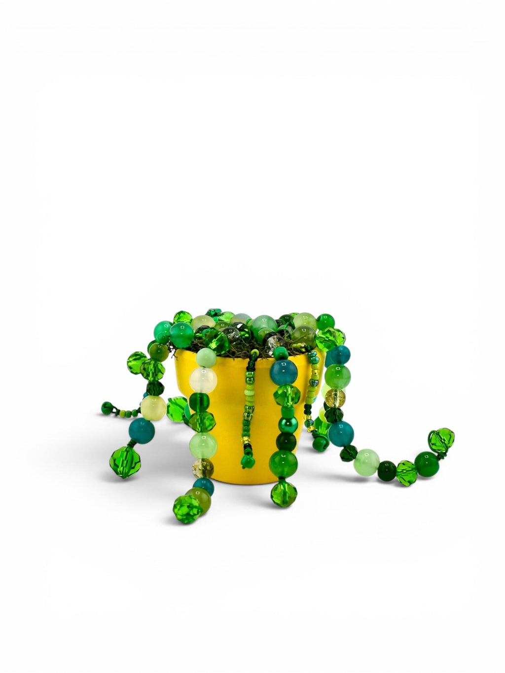 A bright yellow ceramic pot filled with moss and cascading strands of green beads in various shades, sizes, and textures, including translucent, opaque, and faceted glass spheres that hang over the sides of the container.