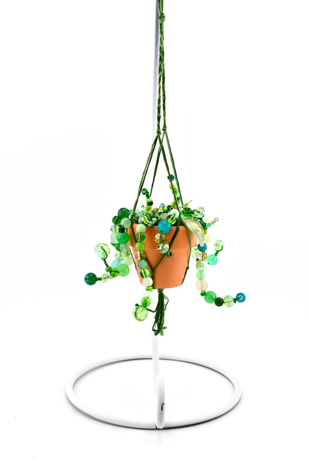 A miniature terracotta pot containing a beaded plant with various shades of green glass beads, suspended by a green braided cord macrame hanger. The entire piece hangs from a white metal stand with a circular base and a curved hook.