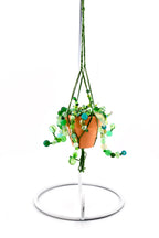 A miniature terracotta pot containing a beaded plant with various shades of green glass beads, suspended by a green braided cord macrame hanger. The entire piece hangs from a white metal stand with a circular base and a curved hook.
