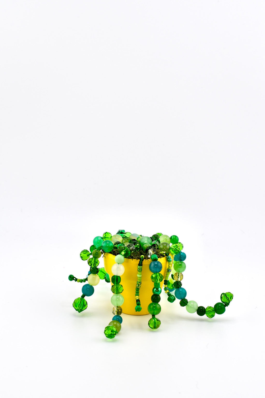A bright yellow pot containing a decorative plant made of various green, lime, and teal glass beads strung together to resemble trailing vines. The beads vary in size, shape, and opacity, creating a textured, cascading appearance.