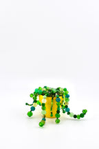 A bright yellow pot containing a decorative plant made of various green, lime, and teal glass beads strung together to resemble trailing vines. The beads vary in size, shape, and opacity, creating a textured, cascading appearance.