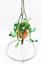 A miniature terracotta pot containing green moss and cascading strands of various green glass and plastic beads. The pot is suspended by dark green braided cord in a macrame style, creating a hanging plant ornament with bead vines.