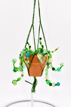 A miniature terracotta pot held in a green macrame hanger, featuring cascading strands of green, teal, and clear glass beads. The beaded vines include various shapes and sizes, with a single translucent leaf-shaped bead accent.