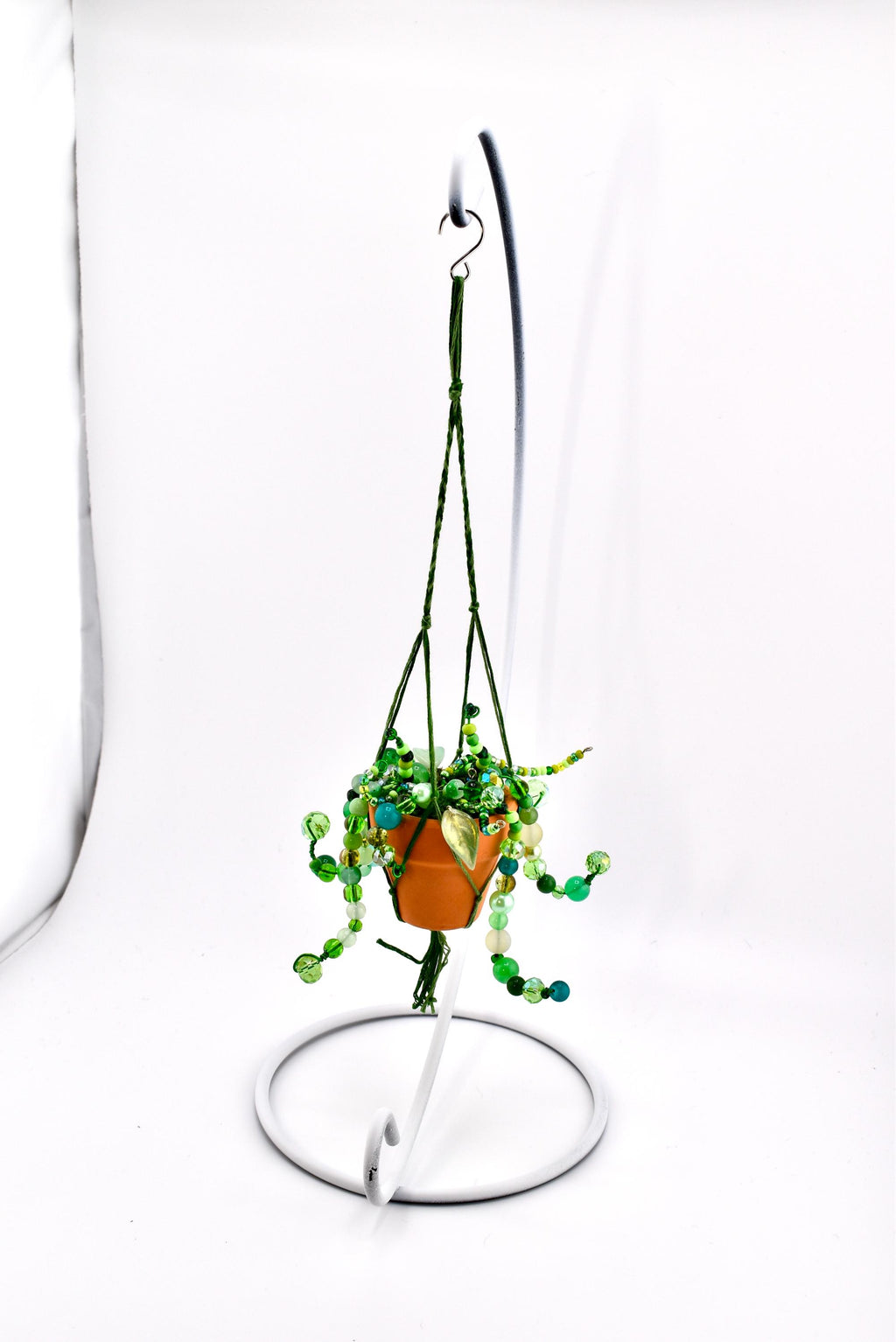 A miniature terracotta-colored pot containing a beaded plant with various shades of green glass beads, suspended by dark green cord in a macrame hanger. The entire piece hangs from a white metal display stand with a circular base.