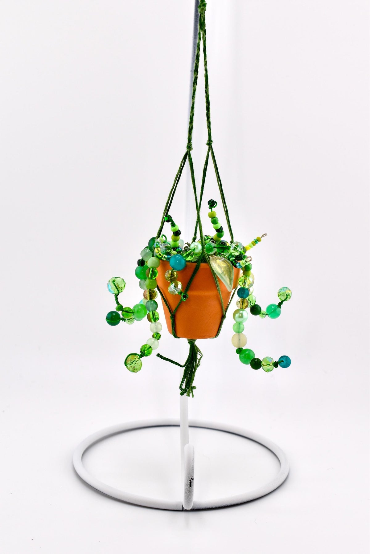 A miniature orange terracotta pot held in a green macrame hanger, featuring cascading strands of green, lime, and teal glass beads that mimic trailing plant vines. The entire piece is suspended from a white metal display stand.