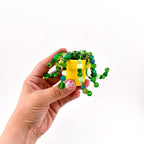 A miniature yellow ceramic pot containing a decorative plant made of various green and translucent beads. The beaded strands hang over the rim of the pot, featuring different shades of green, lime, and teal glass beads in assorted sizes.