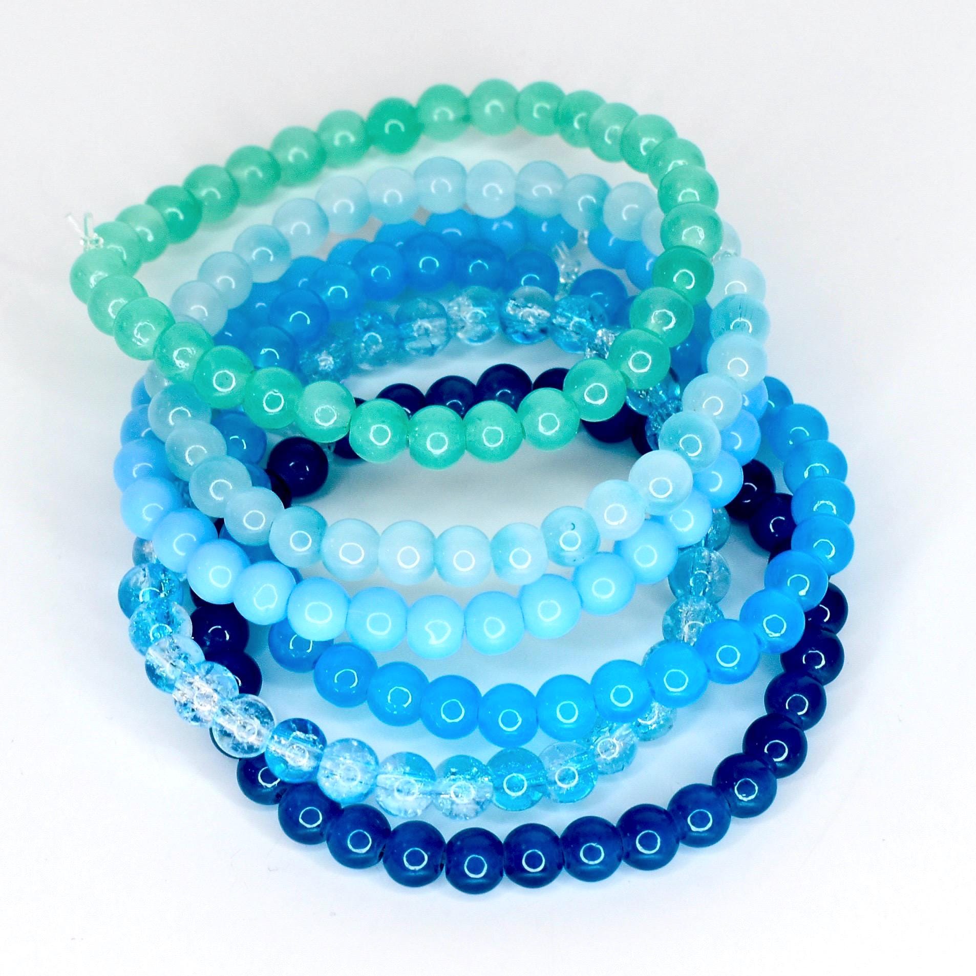 A collection of beaded bracelets in shades of blue and green. The bracelets are made of small, round beads and vary in color from light turquoise to deep navy. The beads are strung together, creating a stackable accessory.
