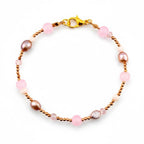 A delicate bracelet featuring a gold-toned chain interspersed with smooth pink spherical beads, small off-white beads, and irregular brown oval-shaped pearls. The piece is secured with a gold-toned lobster claw clasp closure.