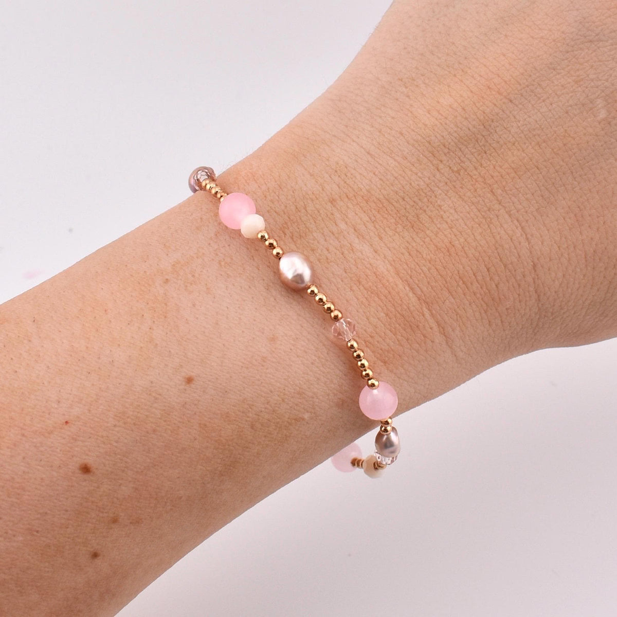 A beaded bracelet featuring a mix of small gold-toned metallic beads, translucent pink spherical beads, and oval-shaped iridescent pearls. The beads are arranged in a repeating pattern along the length of the delicate string.