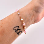 A beaded bracelet featuring a mix of small gold-toned metallic beads, translucent pink spherical beads, iridescent white beads, and smooth oval-shaped pearls. The bracelet is strung on a flexible cord and displays a varied arrangement of these elements.