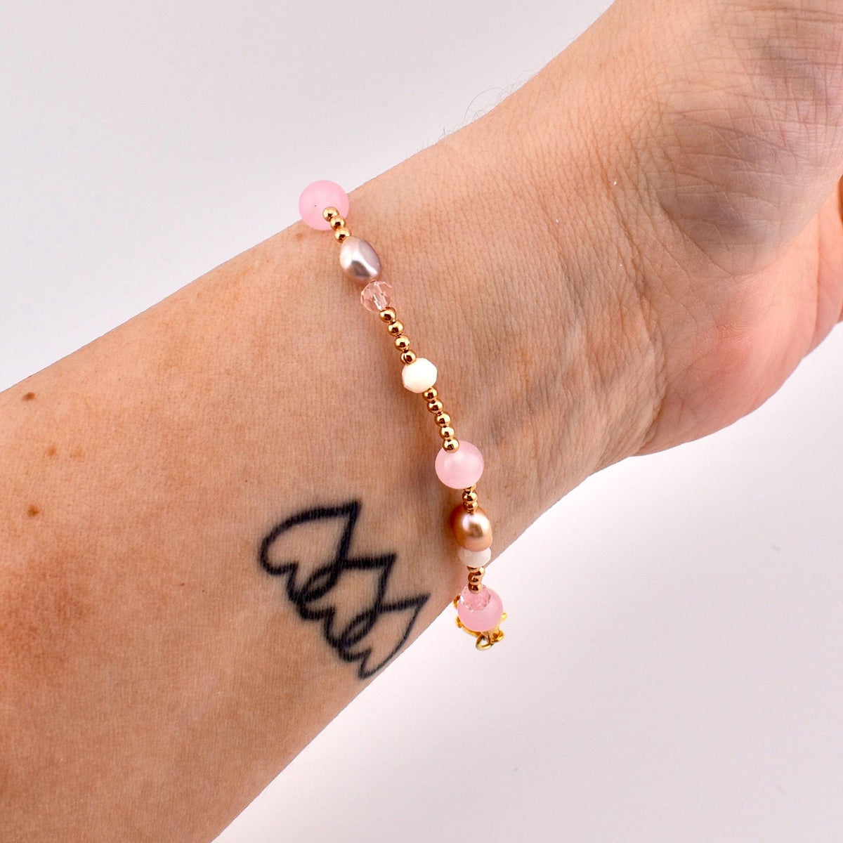 A beaded bracelet featuring a mix of small gold-toned metallic beads, translucent pink spherical beads, iridescent white beads, and smooth oval-shaped pearls. The bracelet is strung on a flexible cord and displays a varied arrangement of these elements.