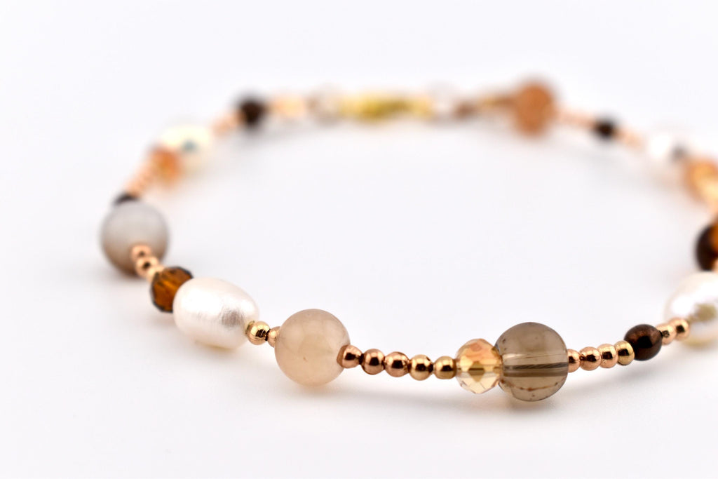 A beaded bracelet featuring a mix of white freshwater pearls, translucent beige and smoky quartz spheres, and small faceted amber-colored crystals, all interspersed with tiny metallic rose gold spacer beads on a thin cord.