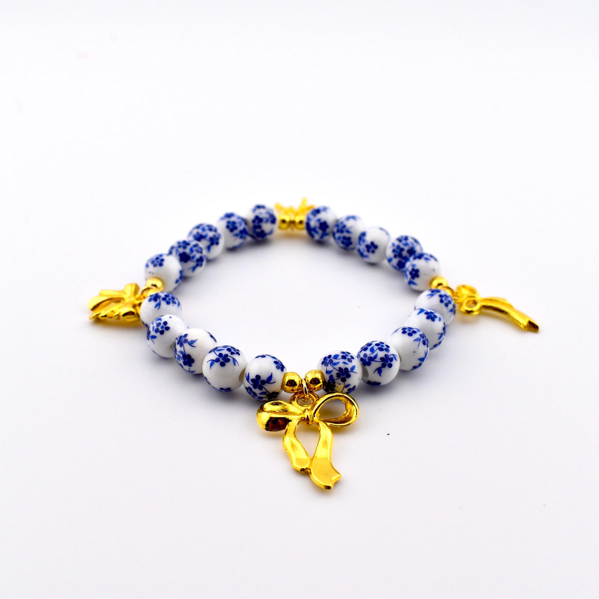 A beaded bracelet featuring round white beads with a blue floral pattern, interspersed with small gold-toned spherical spacers. The bracelet includes three gold-toned bow charms attached at intervals along the circular string of beads.