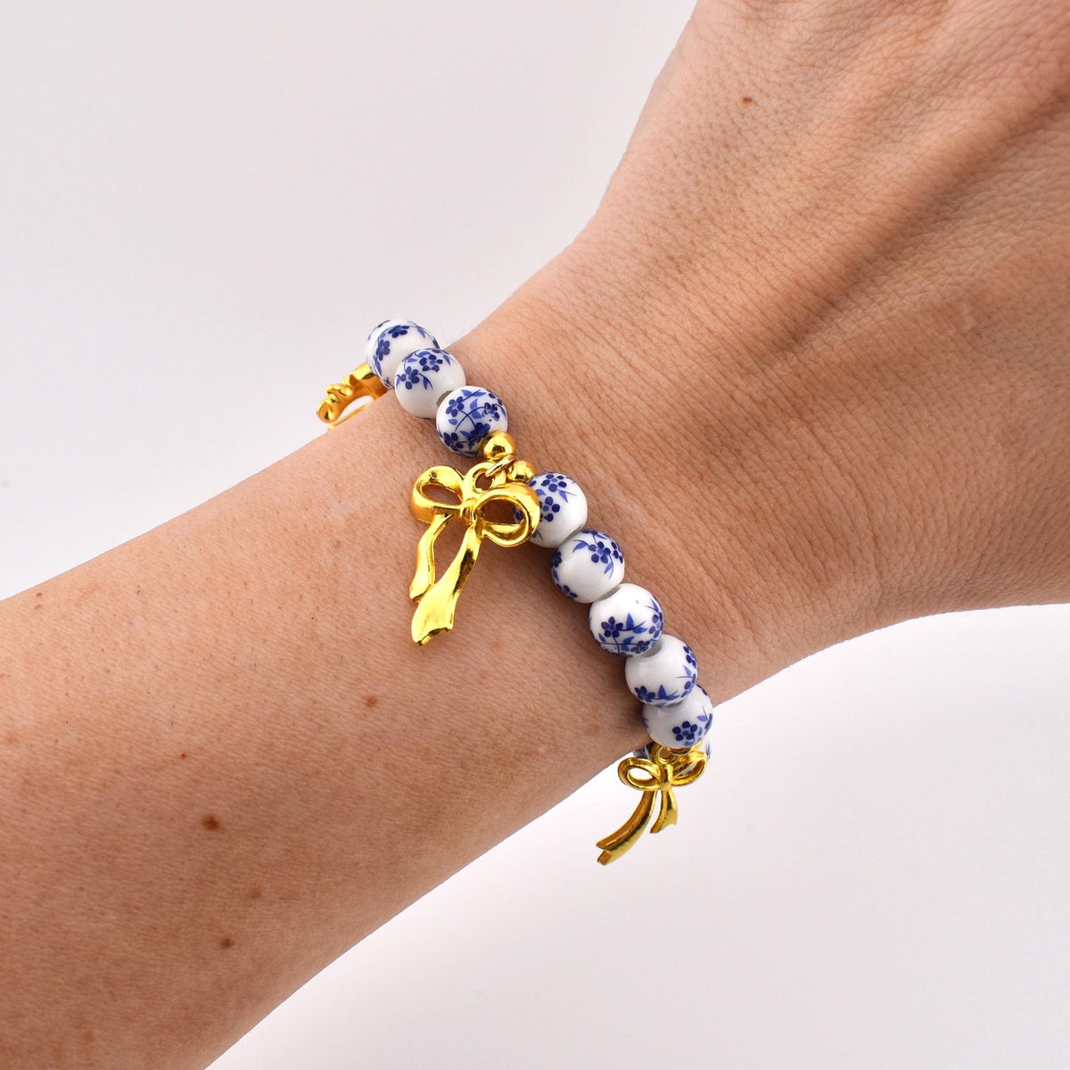 A beaded bracelet featuring round white ceramic beads decorated with a blue floral pattern. The bracelet is accented with two polished gold-tone metal bow charms, one positioned near the center and one at the end of the bead strand.