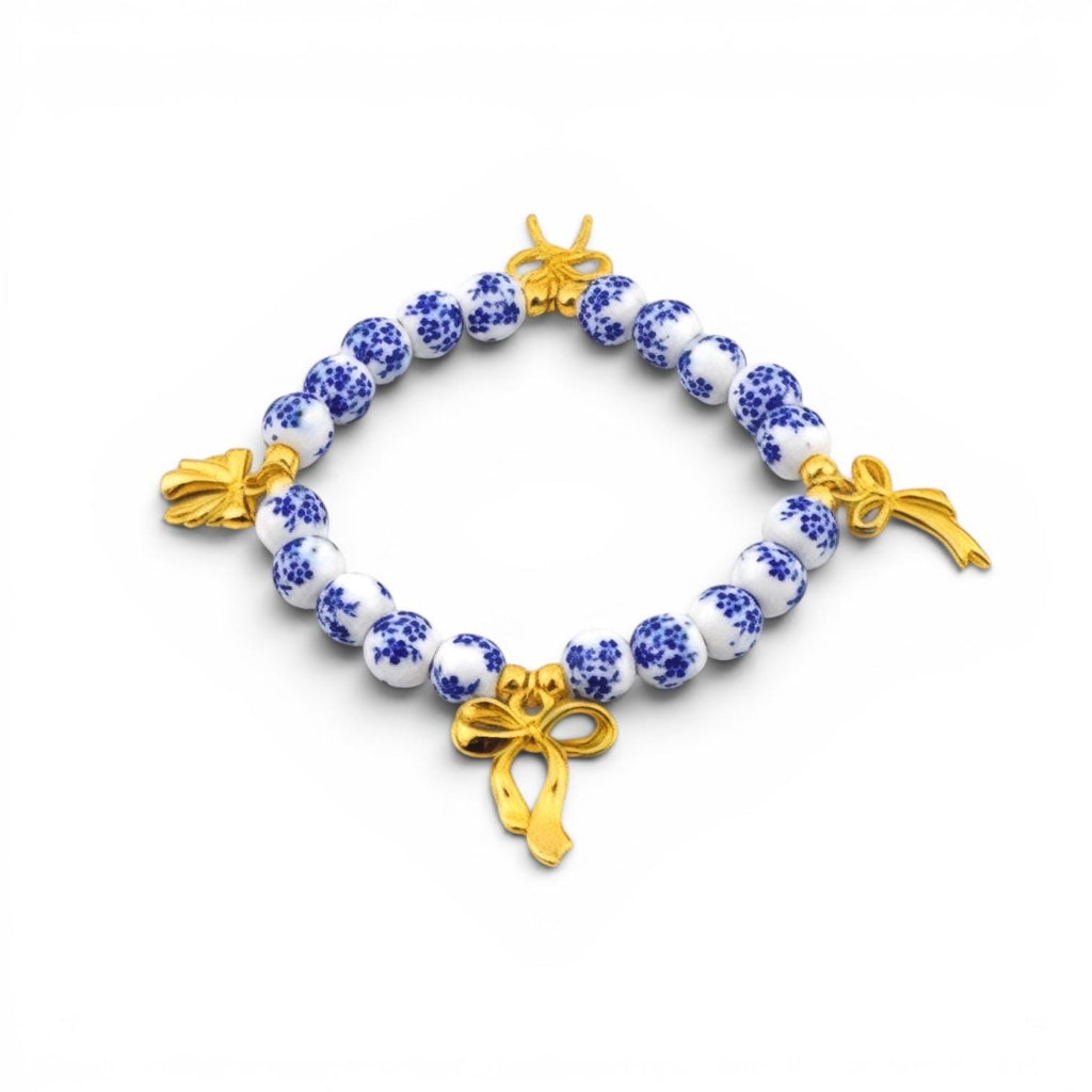 A beaded bracelet featuring round white beads with a blue floral pattern, interspersed with three polished gold-tone metal bow charms. The beads are arranged in a circular shape, creating a contrast between the blue and white ceramic-style beads and the metallic gold accents.