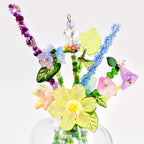 A decorative bouquet of handcrafted beaded flowers featuring a central yellow daffodil, pink and blue bell-shaped blossoms, and purple and blue beaded stems. The arrangement includes green glass leaves and clear crystal accents.