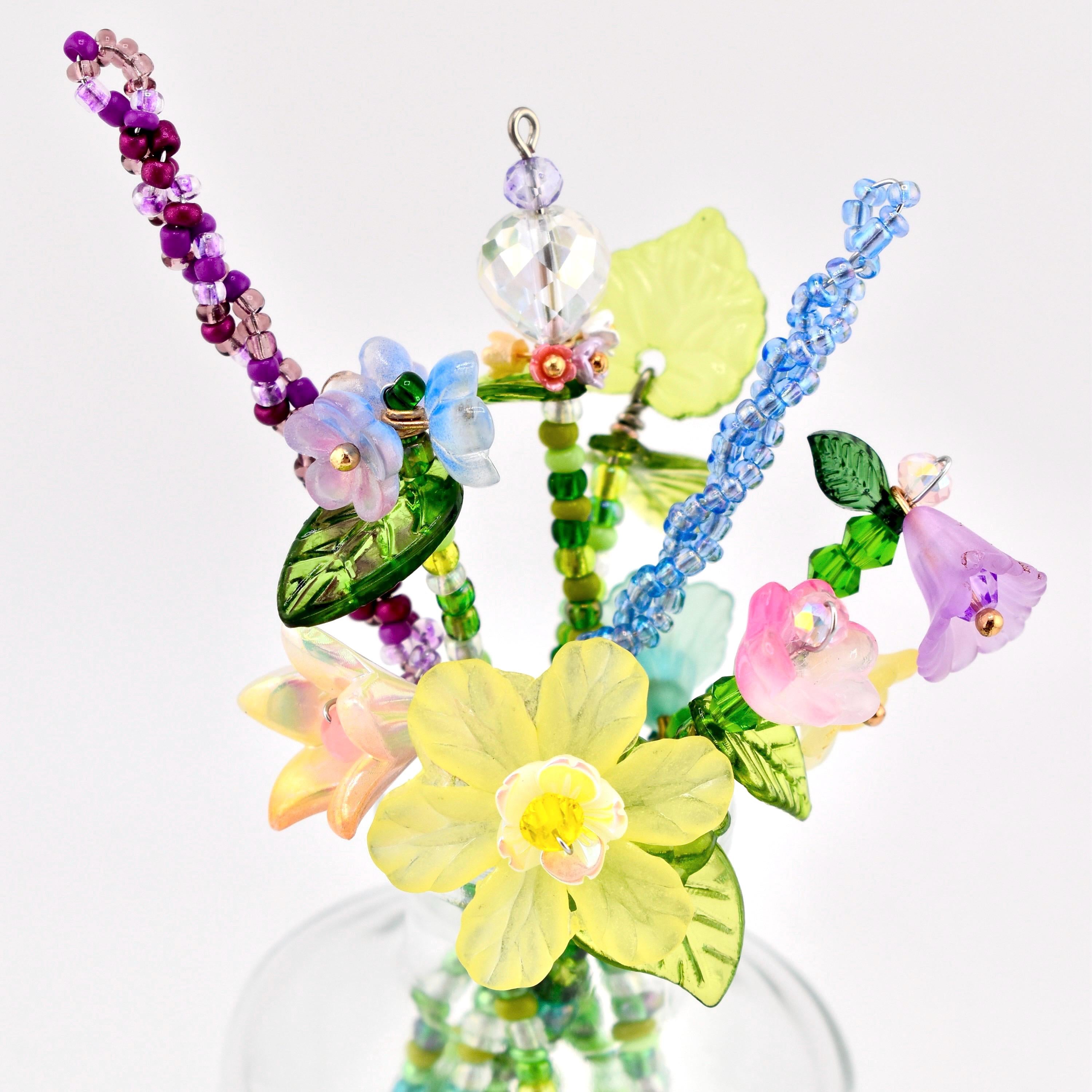 A decorative bouquet of handcrafted beaded flowers featuring a central yellow daffodil, pink and blue bell-shaped blossoms, and purple and blue beaded stems. The arrangement includes green glass leaves and clear crystal accents.