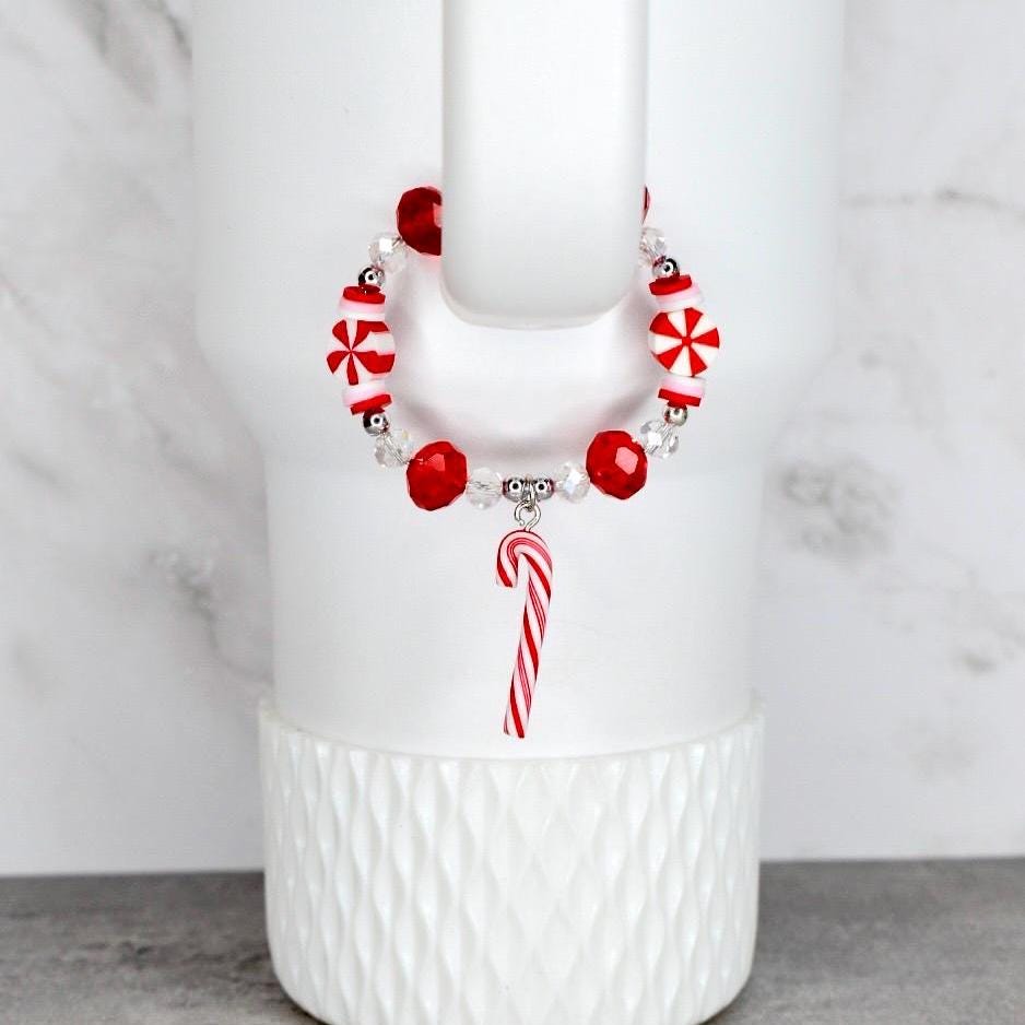 A festive bracelet with red and white candy-themed beads and a candy cane charm. The bracelet is strung on a clear elastic cord. The bracelet is displayed on a white tumbler.