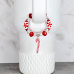 A festive bracelet with red and white candy-themed beads and a candy cane charm. The bracelet is strung on a clear elastic cord. The bracelet is displayed on a white tumbler.