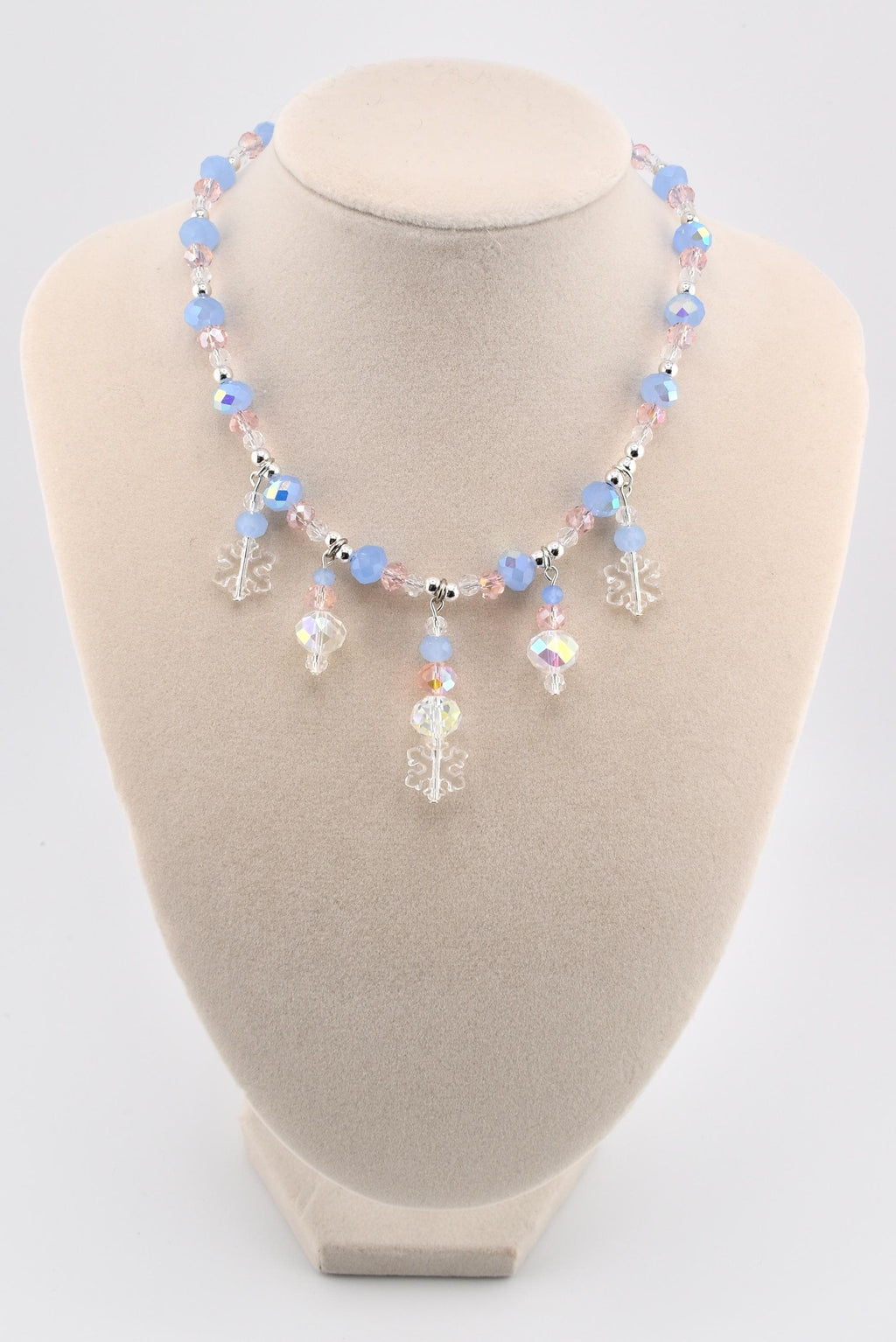 A beaded necklace featuring light blue and pale pink faceted crystal beads interspersed with silver-toned spacers. The necklace includes five dangling charms, consisting of three clear acrylic snowflake shapes and two iridescent crystal drops.