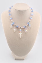 A beaded necklace featuring light blue and pale pink faceted crystal beads interspersed with silver-toned spacers. The necklace includes five dangling charms, consisting of three clear acrylic snowflake shapes and two iridescent crystal drops.