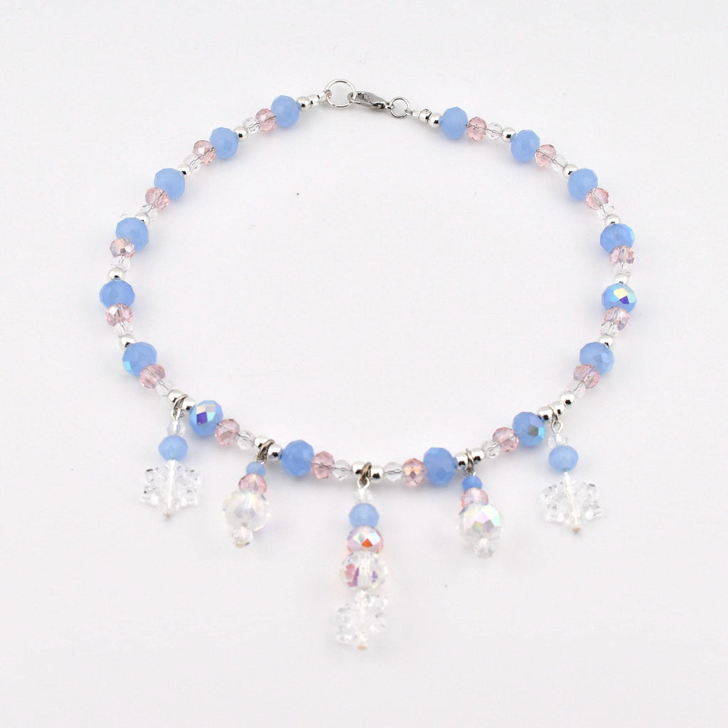 A beaded anklet featuring light blue and pale pink faceted beads, interspersed with small clear crystals. The piece includes five dangling charms, with three clear snowflake-shaped pendants and two round iridescent crystal beads.
