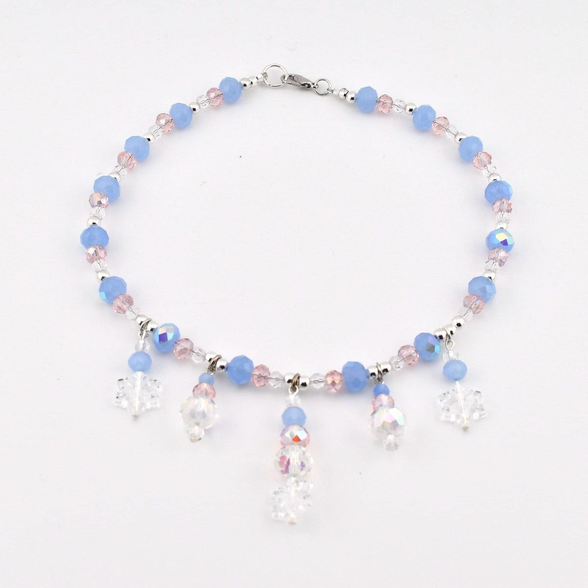 A beaded anklet featuring light blue and pale pink faceted beads, interspersed with small clear crystals. The piece includes five dangling charms, with three clear snowflake-shaped pendants and two round iridescent crystal beads.