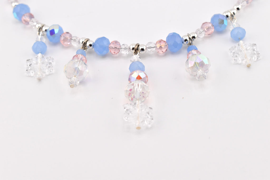A necklace featuring a strand of light blue, pale pink, and clear faceted beads interspersed with silver-toned spacers. Five dangling charms, each composed of a clear snowflake-shaped bead and smaller round beads, hang from the main strand.