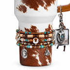 A white tumbler with a brown cowhide print pattern, adorned with a stack of beaded bracelets featuring turquoise, brown, and metallic beads. Charms include a horseshoe, cowboy hat, horse head, and a square star pendant.