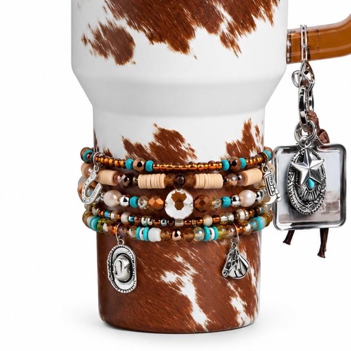 A white tumbler with a brown cowhide print pattern, adorned with a stack of beaded bracelets featuring turquoise, brown, and metallic beads. Charms include a horseshoe, cowboy hat, horse head, and a square star pendant.