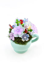 A light mint green ceramic teacup filled with green moss and an arrangement of colorful glass and acrylic flowers. The floral display features various shades of purple, blue, red, and yellow petals with small clear crystal accents.