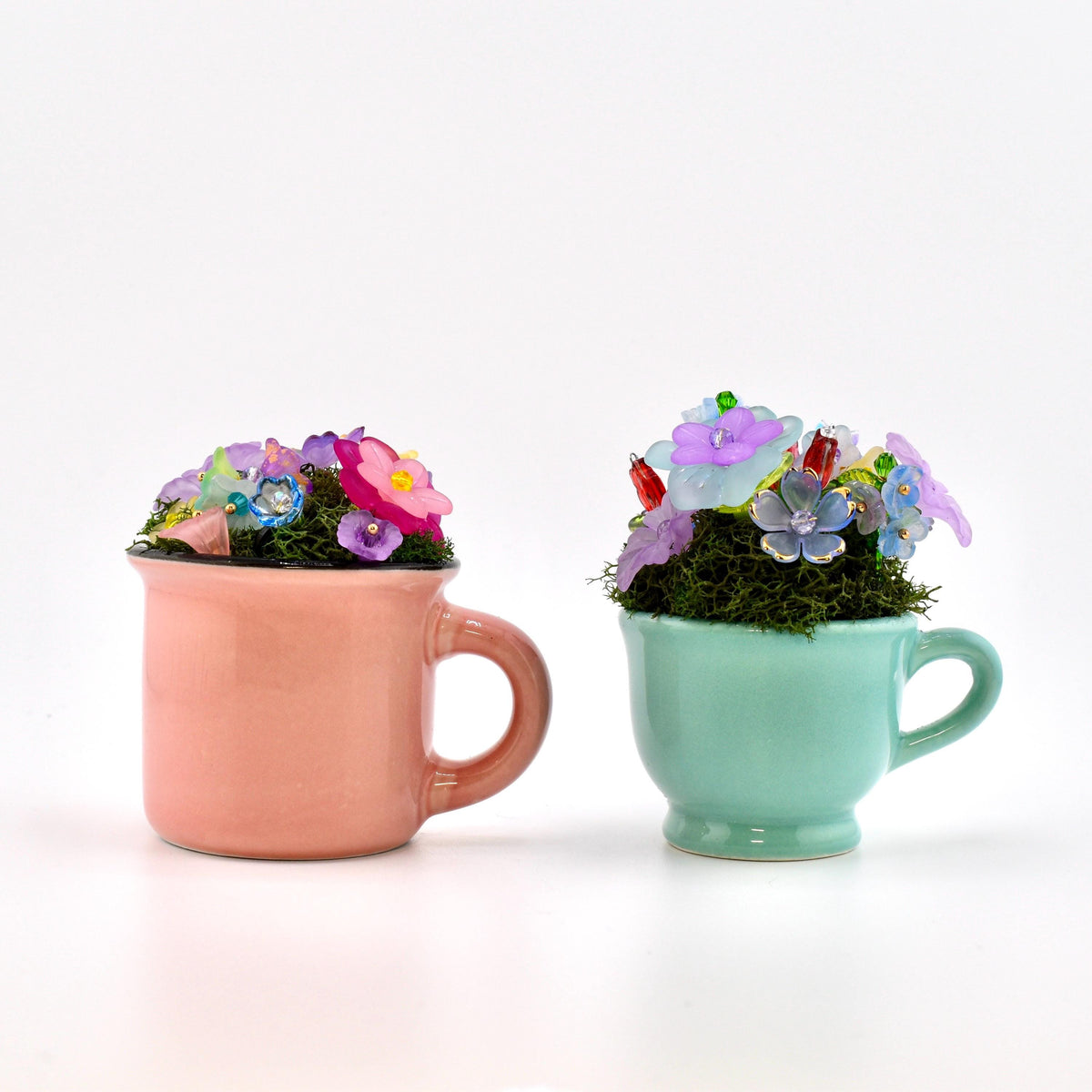Two small ceramic mugs, one peach and one mint green, each filled with dark green moss and an arrangement of colorful, translucent plastic flowers in shades of pink, purple, blue, and red, featuring small bead accents.