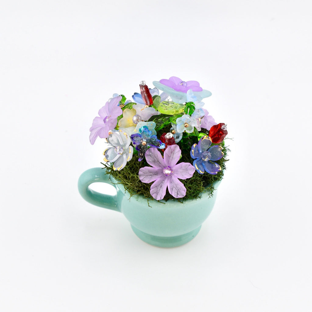 A miniature light blue ceramic teacup filled with green moss and an arrangement of small, multi-colored glass flowers in shades of purple, blue, yellow, and red, accented with clear beads and green glass leaves.