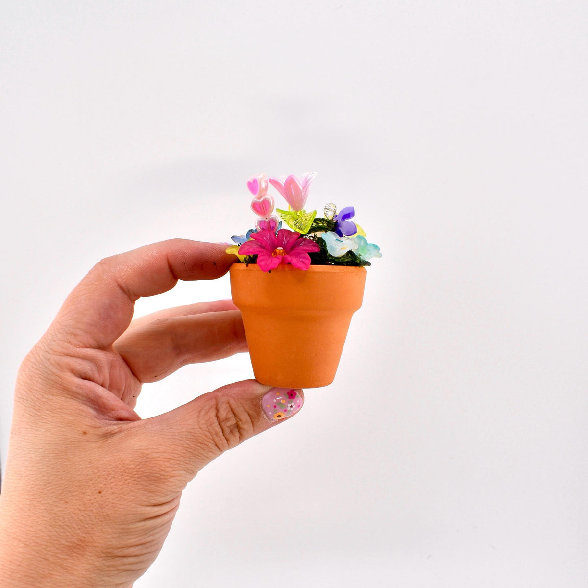 A miniature terracotta-colored clay pot filled with an assortment of small, colorful artificial flowers in shades of pink, purple, blue, and yellow, featuring tiny heart-shaped accents nestled among the vibrant floral arrangement.