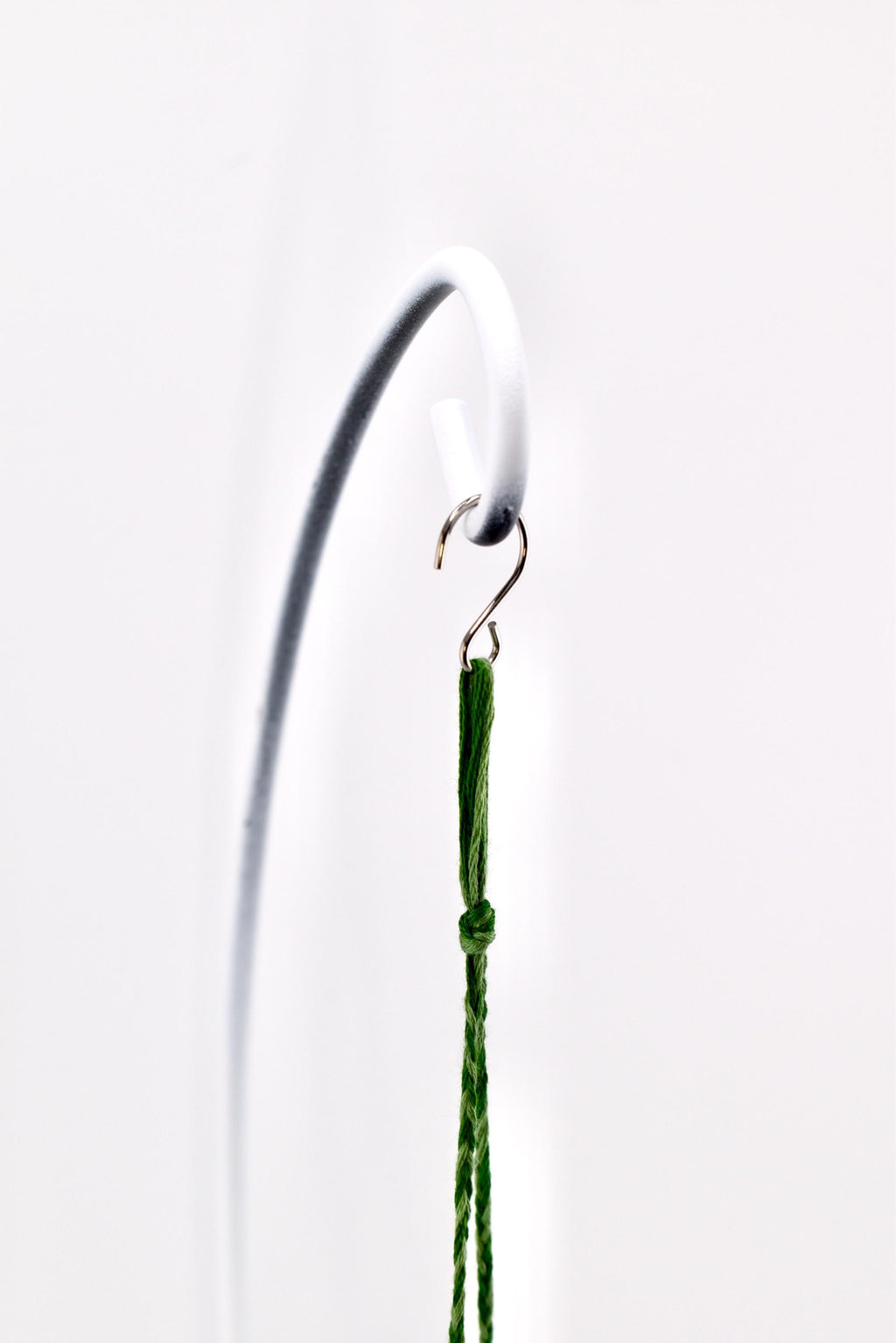 A thin, dark green braided cord featuring a small knot near the top, suspended from a metallic S-shaped hook that is attached to a curved white metal stand. The cord hangs vertically, showcasing its textured, woven fiber construction.