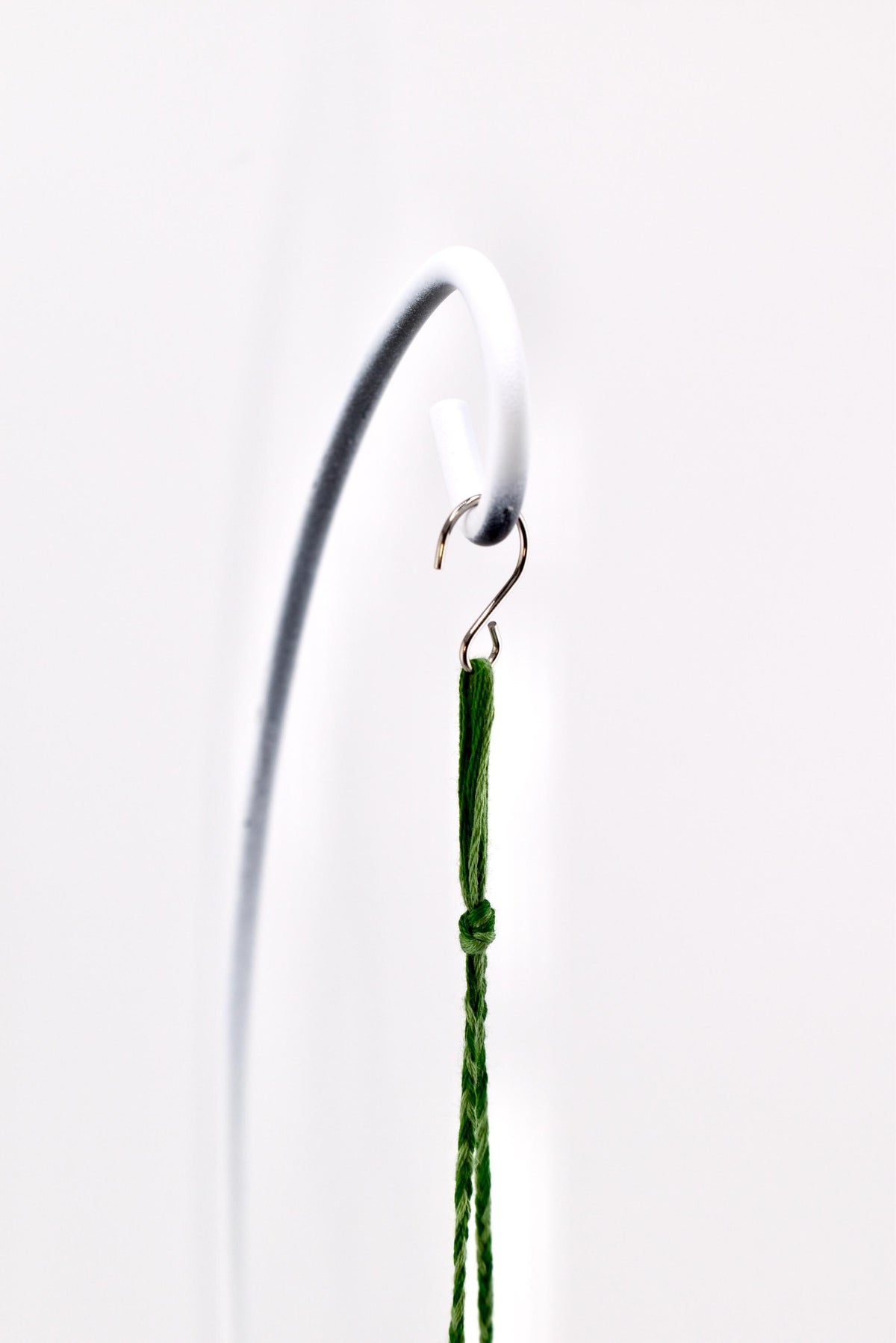 A thin, dark green braided cord featuring a small knot near the top, suspended from a metallic S-shaped hook that is attached to a curved white metal stand. The cord hangs vertically, showcasing its textured, woven fiber construction.