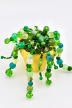A small yellow pot filled with green moss and numerous trailing strands of beads in various shades of green, teal, and lime. The beads are a mix of faceted crystal, smooth round, and translucent glass, all attached to thin green wire.