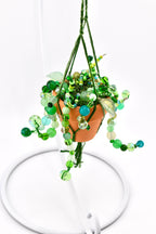 A miniature terracotta pot held in a green macrame hanger, filled with cascading strands of green glass beads in various shades and textures, mimicking a trailing plant. The beads include translucent, opaque, and faceted finishes.