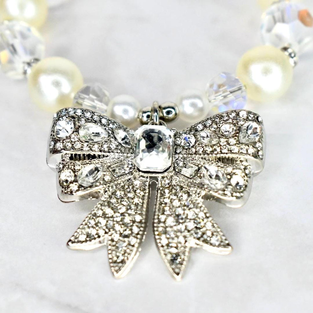 A close-up of a necklace featuring a silver bow pendant encrusted with clear crystals. The bow is the focal point, with a large, rectangular crystal at its center. The necklace includes cream-colored pearls and clear crystal beads.