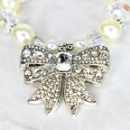 A close-up of a necklace featuring a silver bow pendant encrusted with clear crystals. The bow is the focal point, with a large, rectangular crystal at its center. The necklace includes cream-colored pearls and clear crystal beads.