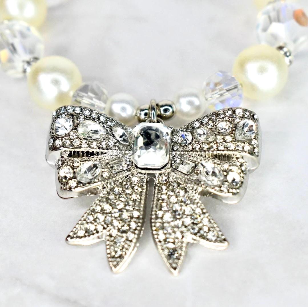 A close-up of a necklace featuring a silver bow pendant encrusted with clear crystals. The bow is the focal point, with a large, rectangular crystal at its center. The necklace includes cream-colored pearls and clear crystal beads.