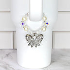 A decorative cup accessory featuring a silver bow adorned with clear rhinestones. The bow is attached to a beaded band with faux pearls and iridescent crystals. The accessory is designed to fit around a white cup.