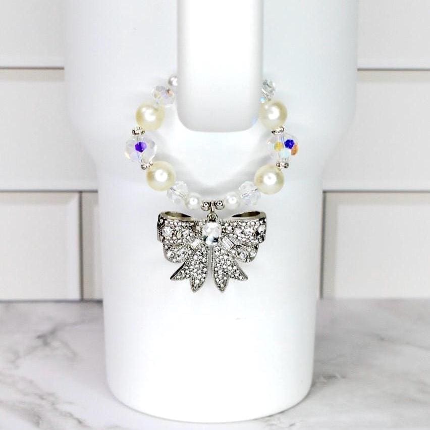 A decorative cup accessory featuring a silver bow adorned with clear rhinestones. The bow is attached to a beaded band with faux pearls and iridescent crystals. The accessory is designed to fit around a white cup.