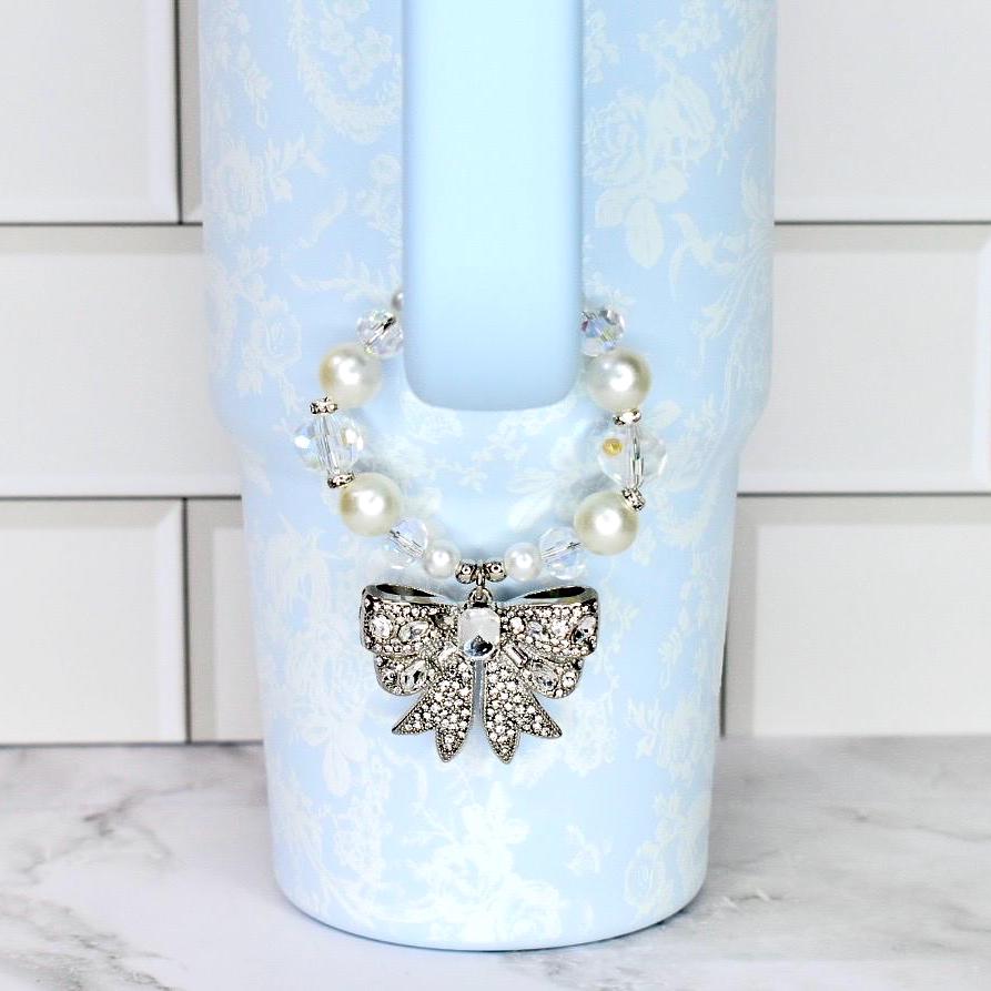 A light blue Stanley with a floral pattern and a decorative bow. The bow is silver with clear rhinestones. The tumbler has a pearl and crystal beaded strap.