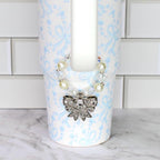 A white tumbler with a light blue floral and bow pattern. A beaded necklace with faux pearls and clear crystals is wrapped around the top. A silver bow pendant with clear rhinestones hangs from the necklace.
