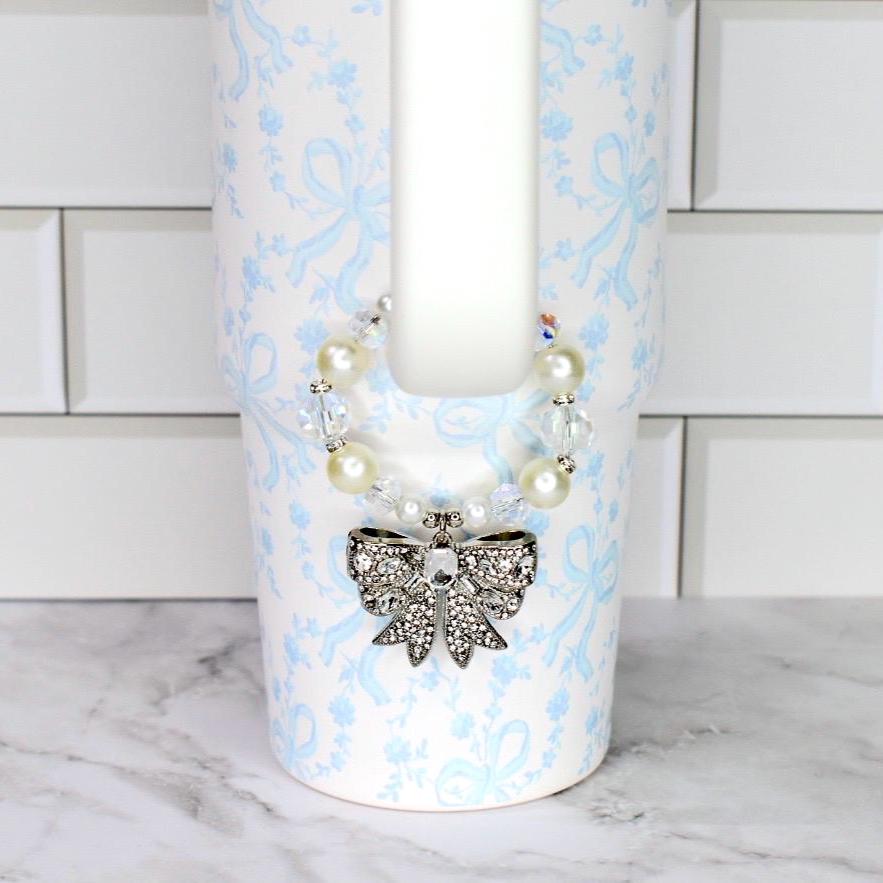 A white tumbler with a light blue floral and bow pattern. A beaded necklace with faux pearls and clear crystals is wrapped around the top. A silver bow pendant with clear rhinestones hangs from the necklace.