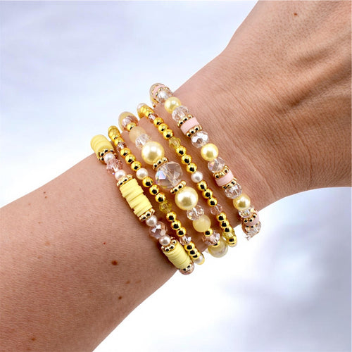 A stack of five bracelets featuring a mix of yellow clay heishi beads, gold-toned metallic spacer beads, clear faceted crystal beads, and smooth pale yellow and soft pink round beads arranged in a layered, multi-textured design.